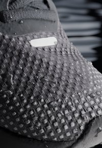 Gray textured athletic shoe upper with a woven design, featuring water droplets and a rubberized white accent near the laces.