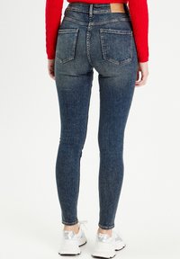 Chude niebieskie dżinsy z wysokim stanem, dwiema tylnymi kieszeniami i kontrastowym przeszyciem. Noszone z białymi butami sportowymi.