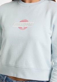 Lichtblauwe sweatshirt met geribde kraag en zoom, met witte en roze geborduurde "Guess Jeans"-logo, gecentreerd binnen een cirkel.
