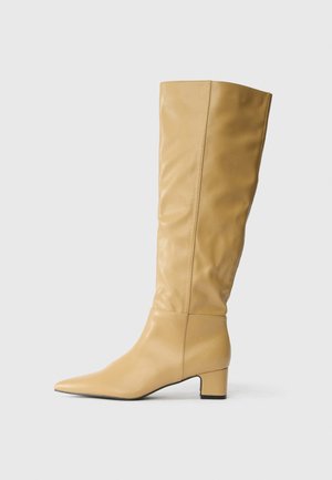 Botte en cuir beige montée jusqu'au genou avec un petit talon carré et un bout pointu, présentée en profil latéral sur un fond blanc.