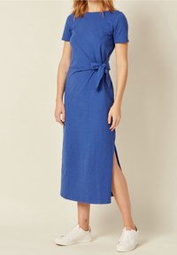 Vestido de algodón azul con mangas cortas, apertura lateral y detalle de cinta en la cintura. Combinado con zapatillas blancas para un look casual.