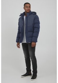 Navy puffer jacket with a textured blue panel, featuring a hood, snap buttons, and two side pockets, paired with dark jeans and black sneakers.