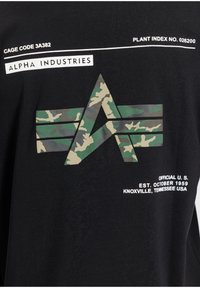 T-shirt noir avec un logo camouflage en forme de "A" et des rayures horizontales, accompagné de texte blanc précisant la marque et l'origine.