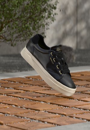 Sneaker in pelle nera con suola bianca e occhielli dorati che fluttua sopra un pavimento in legno all'aperto vicino a una pianta verde sfocata.