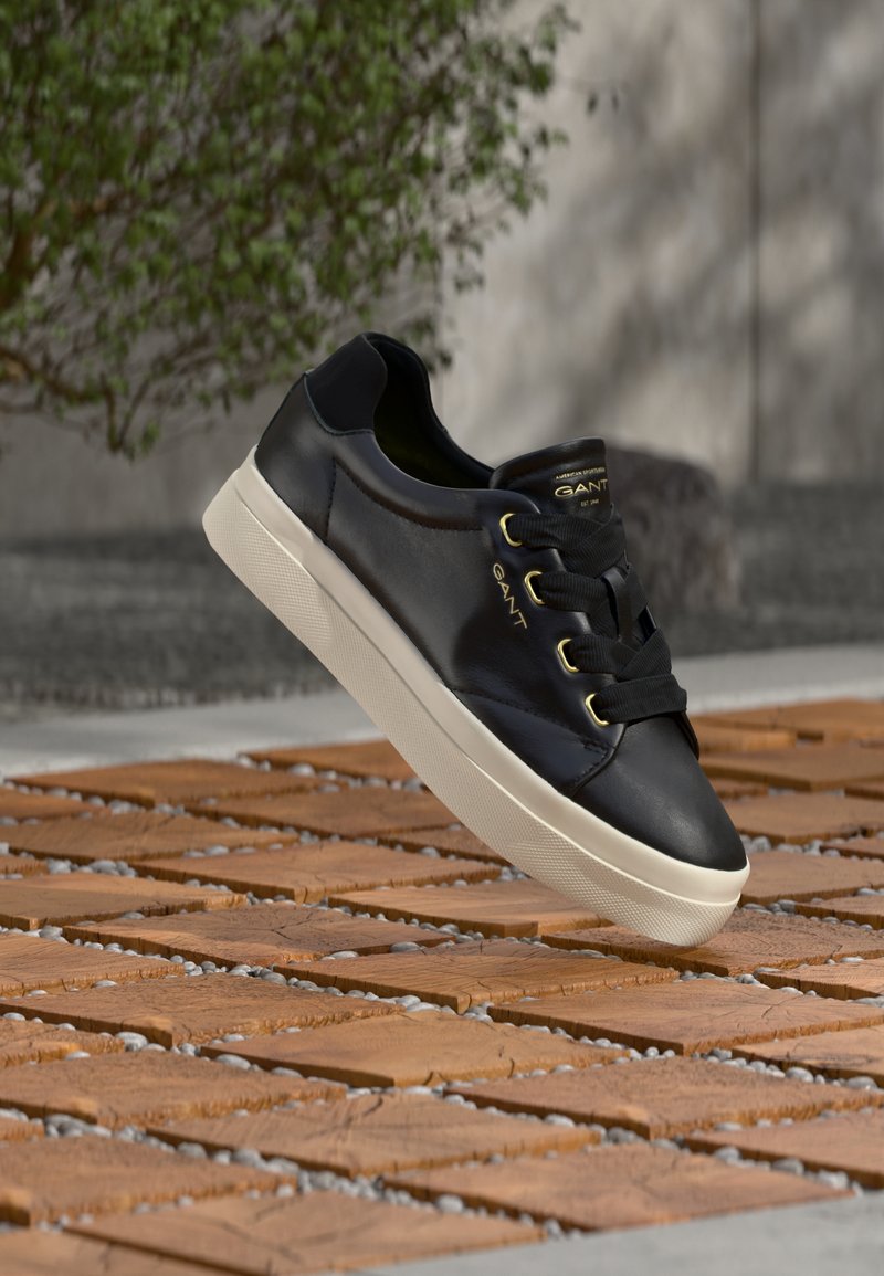 Czarny skórzany trampek z białą podeszwą i złotymi przelotkami unoszący się nad drewnianą płytką na zewnątrz, w pobliżu rozmytej zielonej rośliny.