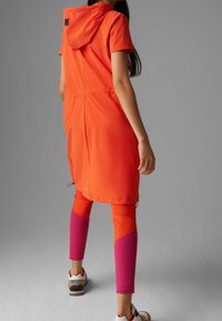 Giacca antipioggia arancione con cappuccio, maniche corte e vita con cordoncino; abbinata a leggings con pannelli arancioni e rosa; scarpe sportive.
