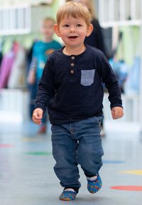 Kind in een marineblauwe henley-shirt met lange mouwen, houten knopen en een lichtblauwe zak, gecombineerd met blauwe jeans en met patronen versierde instappers.