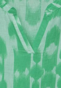 Zelená tunika s výstřhem do V, vyrobená z texturované látky s abstraktním vzorem v různých odstínech zelené. Detaily zahrnují precizní šití.