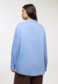Jasnoniebieska, długosleeve koszula z gładkiego materiału. Posiada klasyczny kołnierzyk, mankiety na guziki oraz luźny krój, widok z tyłu.