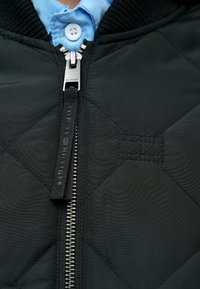 Veste matelassée noire avec fermeture éclair argentée dotée d'une tirette à logo, col côtelé et chemise bleue superposée en dessous.