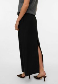 Jupe maxi noire cannelée avec une fente haute, fabriquée en tissu doux. Associée à des talons ouverts marron ornés de œillets décoratifs.