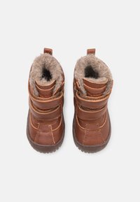 Dvojice hnědých dětských zimních bot s chlupatou šedou podšívkou a dvěma nastavitelnými pásky, pohled shora na bílém pozadí.