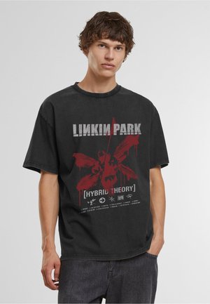 Jonge man draagt een zwart Linkin Park Hybrid Theory T-shirt en donkere spijkerbroek, staand tegen een effen lichte achtergrond.