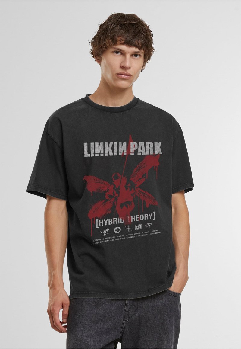 Jeune homme portant un t-shirt noir Linkin Park Hybrid Theory et un jean foncé, debout devant un fond clair uni.