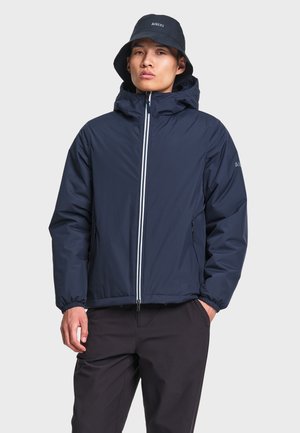 Homme portant une veste à capuche bleu marine avec fermeture éclair frontale et un chapeau bob assorti, debout, une main dans la poche, sur un fond uni.