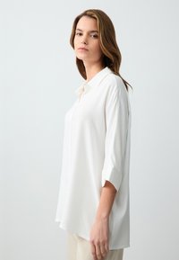 Weiße, gebuttonte Bluse mit Kragen, Dreiviertelärmeln und lockerer Passform; glattes Material mit minimaler Textur und klaren Linien.