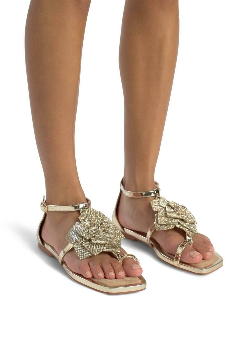 Gouden platte sandalen kenmerken zich door een opvallende, getextureerde bloemaccent, een enkelbandje met gesp en een minimalistisch ontwerp dat de open tenen beklemtoont.