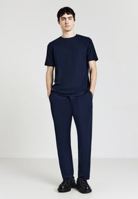 Giovane uomo in piedi con le mani in tasca, indossa una maglietta blu navy, pantaloni abbinati e scarpe nere su uno sfondo chiaro e semplice.