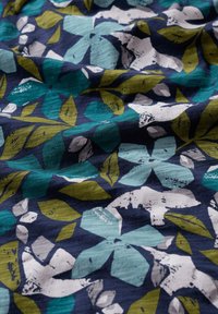 Kwiatowy materiał z dużymi, nachodzącymi na siebie kwiatami w odcieniach turkusu, bieli i zieleni na ciemnoniebieskim tle; gładka tekstura.