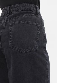 Svarta denimjeans med hög midja, vida ben och bakficka med stickad detalj. Tyget har en mjuk textur med subtila blekningar.