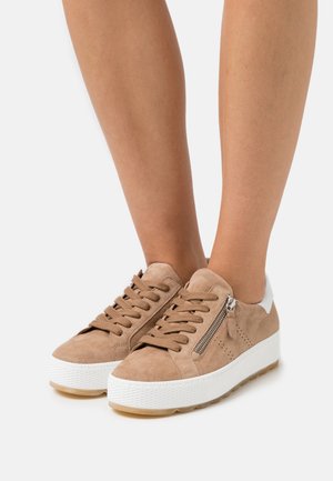 Beige suede sneakers with white rubber sole, featuring lace-up front and side zip detail. Textured upper with perforations for breathability.