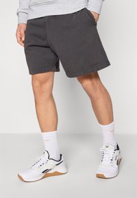 Σκούρο γκρι βαμβακερά σορτς με ελαστική μέση, πλαϊνές τσέπες και εμφανείς ραφές, σε συνδυασμό με λευκά αθλητικά παπούτσια και κάλτσες.