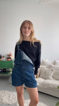 Jeune femme aux cheveux blonds portant un top noir à manches longues et une salopette courte en jean, debout dans un salon lumineux avec un canapé blanc et une armoire verte.