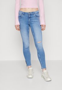 Vaqueros ajustados de color azul claro con detalles desgastados en la rodilla izquierda, que cuentan con un diseño clásico de cinco bolsillos y cremalleras en los tobillos.