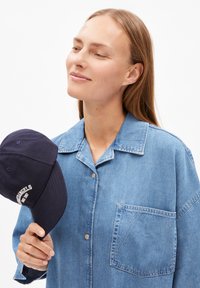 Blauwe denim blouse met een kraag en zak. Houdt een marineblauwe pet vast met witte tekst. Soepele textuur, casual ontwerp en relaxte pasvorm.