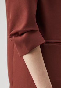 Maroon tunic with a textured fabric. Features a rolled cuff and a sleek design, highlighting subtle stitching and a side pocket.