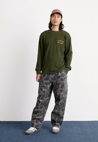 Zielona koszulka z długim rękawem w splot wafel, z żółtą grafiką, szare spodnie cargo w camo oraz szare sneakersy na niebieskim wykładzinie.