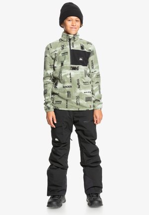 Pull en polaire vert à motifs avec une poche poitrine noire ; associé à un pantalon de neige noir. Le mannequin porte un bonnet noir et des bottes.