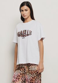 T-shirt in cotone bianco con stampa in grassetto "GAELLE paris" in lettere con stampa animale testurizzata. Vestibilità ampia con scollo rotondo e maniche corte.