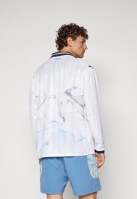 Langärmliges, weißes Hemd mit blauem Aquarell-Mountain-Print auf dem Rücken, horizontalen Streifen, navyfarbenem Kragen und leichtem Stoff.