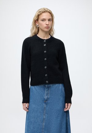 Femme blonde portant un cardigan noir boutonné, des boucles d'oreilles en argent et une jupe en jean bleue, debout devant un fond clair uni.