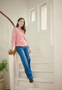Rosa Pullover mit Zopfmuster, blaue Jeans mit geradem Bein und dunkle Schuhe. Steht auf einer Treppe mit Holzgeländer und weißen Wänden; kleine Pflanze in der Nähe.