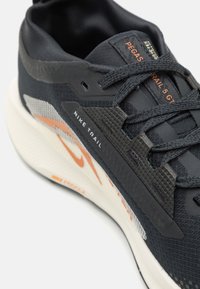 Chaussure de trail Nike noire avec une maille grise et des accents orange. Elle présente une tige texturée, un col rembourré et une semelle blanche amortissante.