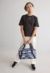 Puma tornazsák kék és fehér grafikával, piros és fekete részletekkel, masszív fekete fogantyúkkal, egy fekete pólót viselő személy kezében.