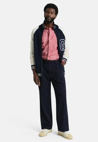 Marineblauwe en crèmekleurige gebreide varsityjack met "G"-logo, lichtroze button-up overhemd, marineblauwe broek en crèmekleurige sneakers met gestructureerde zolen.