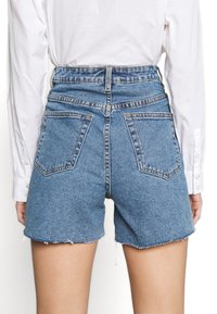 Pantalones cortos vaqueros de talle alto en azul medio con dobladillo deshilachado, que cuentan con dos bolsillos traseros y trabillas para el cinturón. Detalles de costuras visibles por todo el tejido.