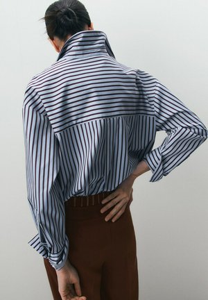 Persona con cabello oscuro recogido en un moño, vestida con una camisa de rayas azules y blancas y pantalones marrones, vista desde atrás con una mano en la cadera.