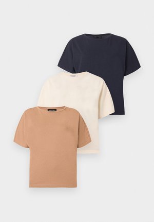 Femme portant un haut beige à manches courtes, un jean bleu à jambes larges, des talons blancs pointus et des lunettes de soleil foncées, debout devant un fond blanc uni.