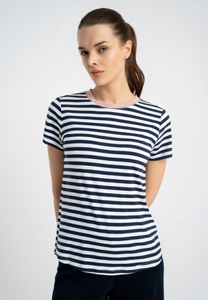 Kobieta nosząca granatowo-biały koszulkę z poziomymi pasiami i różowym kołnierzykiem, stojąca na jednolitym jasnym tle.