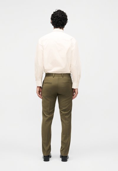 Άνδρας που στέκεται με την πλάτη, φοράει μια εκρού μπλούζα με μακριά μανίκια, ελαιδί παντελόνι και μαύρα επίσημα παπούτσια σε απλό φόντο.