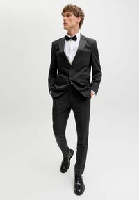 Mladić u crnom fraku s leptir mašnom, bijelom košuljom i crnim svečanim cipelama, stoji s rukama u džepovima, gledajući u stranu.