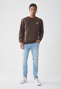 Man in een bruine sweater met een leeuwlogo, lichte spijkerbroek en witte sneakers, staande tegen een effen lichte achtergrond.