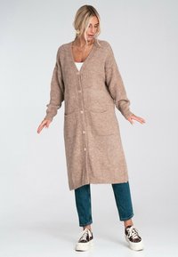 Kobieta ubrana w długi beżowy kardigan na guziki, niebieskie dżinsy i wzorzyste trampki, stojąca na tle jasnego, jednolitego tła.