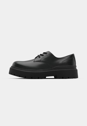 Black leather oxford shoe with a rounded toe, featuring a thick rubber sole and traditional lace-up design. Soft texture, minimal stitching.