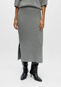 Jupe grise tricotée avec une texture côtelée, mi-longue et fendue sur le côté, assortie à des bottines noires à talons aiguilles. Le pull a un design similaire.