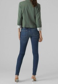 Veste courte verte avec des poignets blancs retroussés, associée à un jean skinny bleu foncé. Le modèle porte des sandales à talons argentées, mettant en valeur une silhouette ajustée.